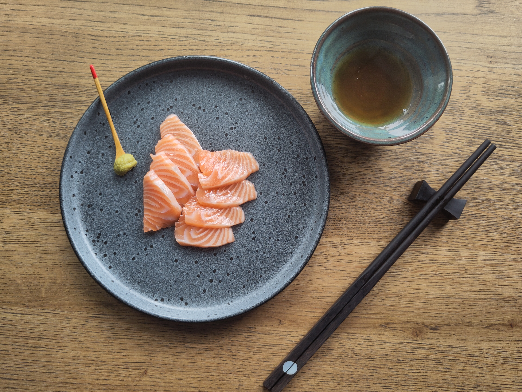 Salmon sashimi, wasabi, chopsticks and a bowl of oat shoyu on an oak table